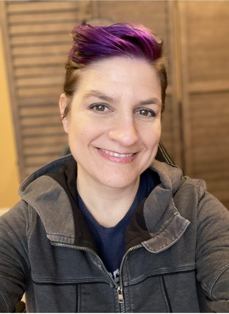 headshot of jennifer cranston. a white woman wearing a gray hooded sweatshirt. her hair is colored purple, short on the sides and is longer on top. she is smiling at the camera. 