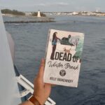 a person's hand is holding Dead of Winter Break paperback on a cruise ship deck with a lighthouse in the background