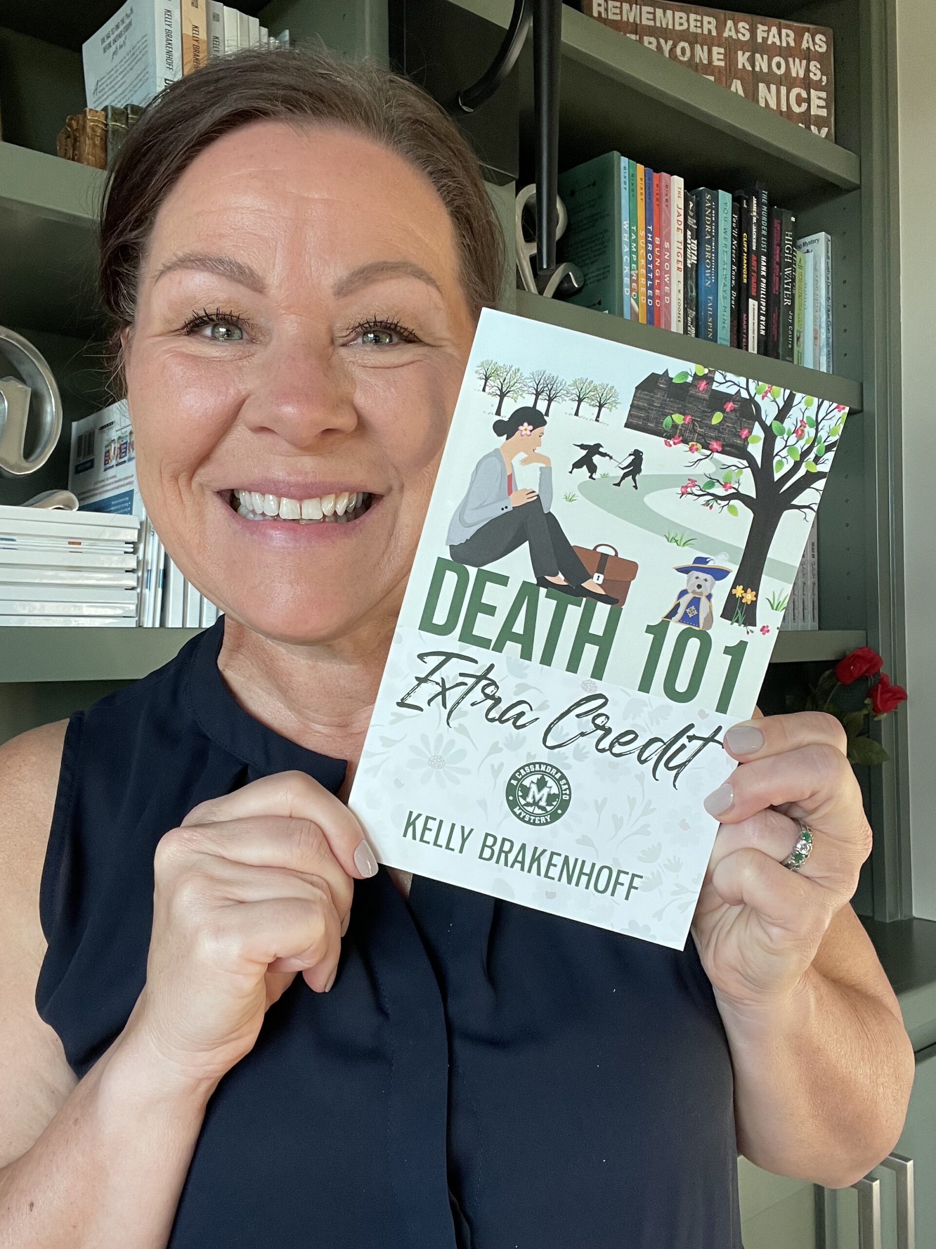 a woman with a dark ponytails holds a paperback copy of the book Death 101: Extra Credit. She is smiling.